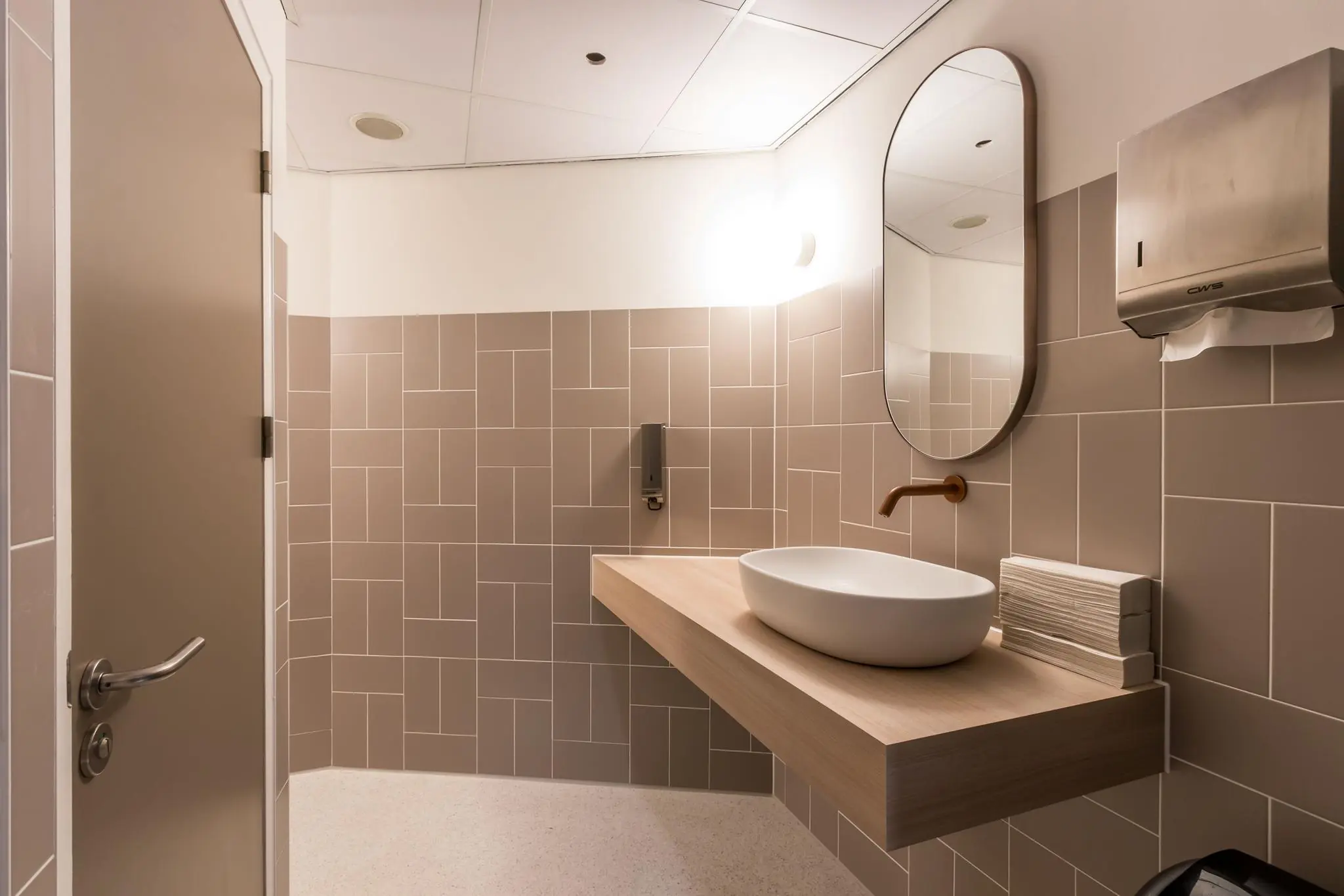 Moderne sanitaire ruimte aan de Vliegend Hertlaan met een ovale spiegel, waskom en beige wandtegels.